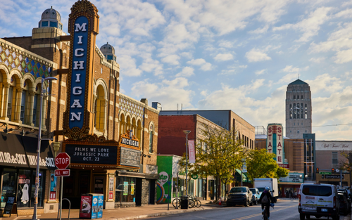 Historic downtown street with storefronts, a tall vertical “Ann Arbor, MICHIGAN” sign, parked cars and a motorbike, and a distant clocktower building under a partly cloudy sky.
